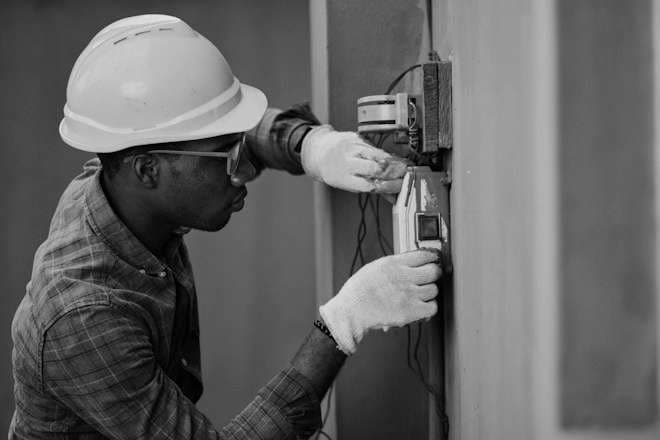 An electrician in a white hard hat and safety glasses tests wiring at a wall panel