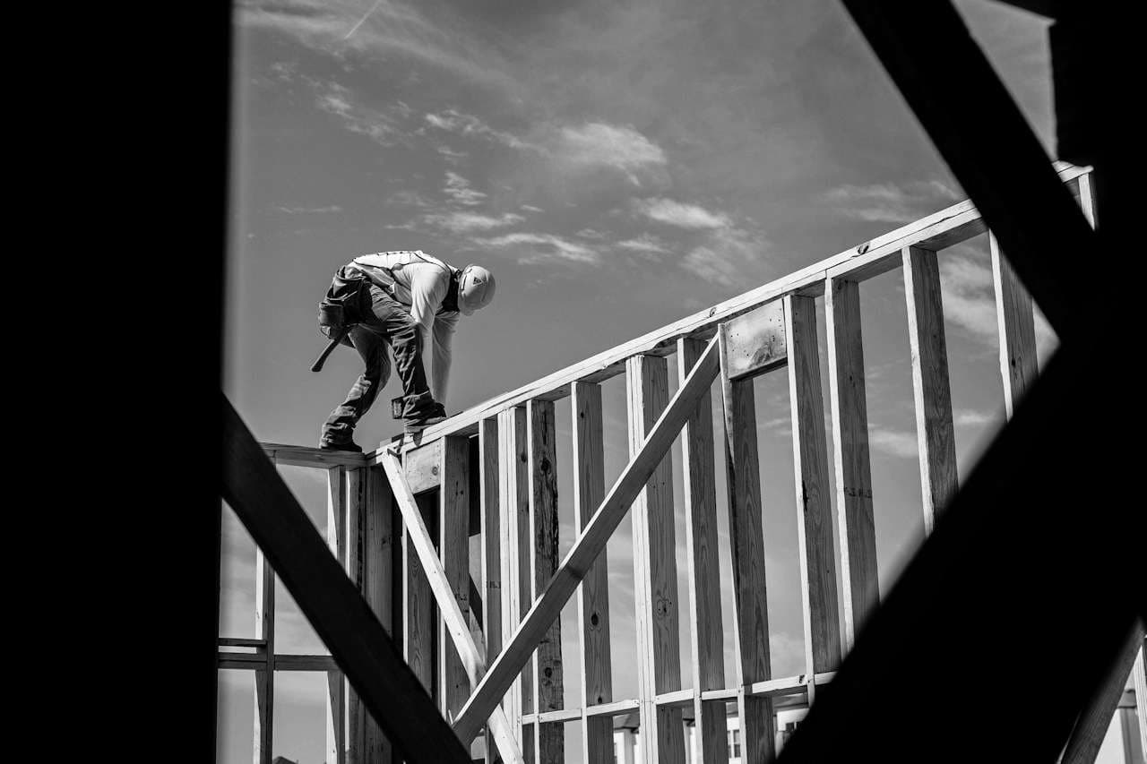 A framer in a hard hat balances on the top plate of a wood-framed wall against the sky